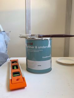 a can of paint next to an orange tool on a white table with a bed in the background