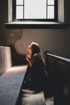 a woman sitting in front of a window with her hands clasped to her chest and looking up