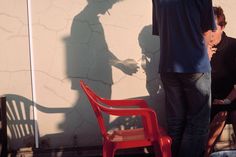 a man standing next to a red chair in front of a wall with a shadow on it