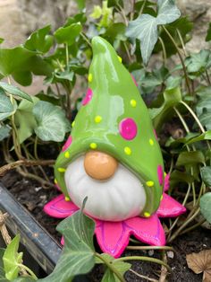 a green and pink gnome statue sitting in the ground next to some ivys with yellow dots on its face