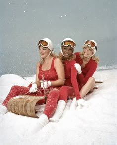 three women in red snowsuits and ski goggles sitting on the snow with their arms around each other