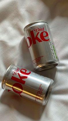 two cans of diet coke sitting on top of a white bed sheet next to each other