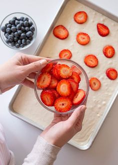 a person holding a bowl with strawberries in it and blueberries on the side