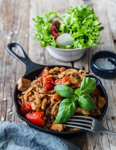 a skillet filled with meat and vegetables on top of a wooden table next to a bowl of salad