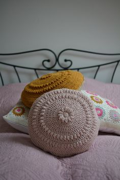 three crocheted pillows sitting on top of a pink bedspread with a metal headboard