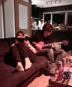 two people sitting on a couch playing guitar and drinking water in the living room at night