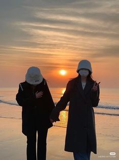 two people walking on the beach holding hands with the sun setting in the distance behind them