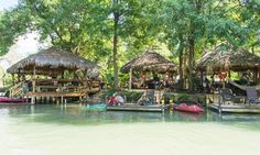 some people are sitting on the dock in front of thatched huts and canoes