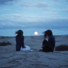 two people sitting in the sand at sunset with their hair blowing in the wind and one person holding a surfboard