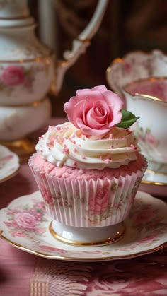 a cupcake on a saucer with a pink rose on top and tea cups in the background