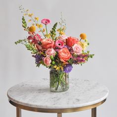 A glass vase with a medium flower arrangement sits on a marble table in front of a white backdrop. The flowers are many colors and include roses Multicolor Floral Arrangement, Bridal Shower Color Schemes Summer, Flower Arrangements For Table Birthday, Artificial Wildflower Arrangements, Live Flower Wedding Bouquets, Wildflower Bouquet In Vase, Bouquet Of Flowers Colorful, Everyday Floral Arrangements, March Floral Arrangements