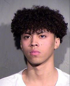 a young man with curly hair wearing a white t - shirt looking at the camera