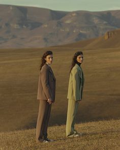 two women standing on top of a dry grass covered field next to each other in front of mountains