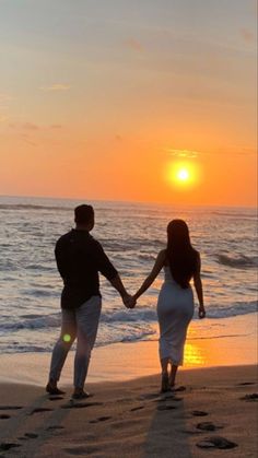 a man and woman holding hands while walking on the beach at sunset with footprints in the sand