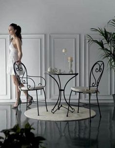 a woman standing in front of a table with two chairs and a vase on it