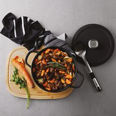 a wooden cutting board topped with a pot filled with stew and carrots next to a spoon