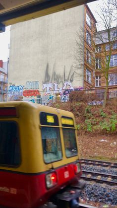 a yellow and red train traveling down tracks near a tall building with graffiti on it