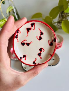 a hand holding a small bowl with red and black designs on it, in front of a plant