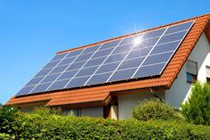 a house with a solar panel on the roof and bushes around it, in front of a blue sky