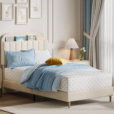 a white bed with blue sheets and pillows in a bedroom next to a lamp on a table