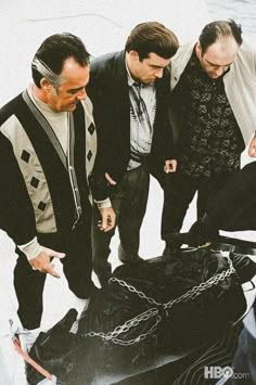 three men standing around a black suitcase with chains on the front and back sides, looking down at it