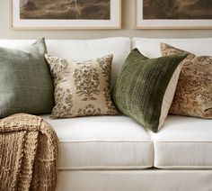 a white couch with green and brown pillows on it's backrest, in front of two framed pictures