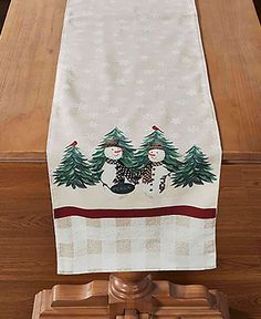 a christmas table runner with two snowmen on it and trees in the background, sitting on a wooden stand