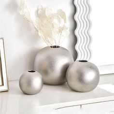 three silver vases sitting on top of a white counter next to a mirror and framed photograph