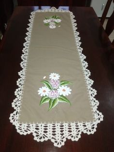 an embroidered table runner with white flowers and green leaves on the border, sitting on a wooden table