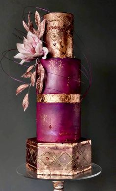 a three tiered purple and gold wedding cake with flowers on top, sitting on a glass plate