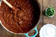 a pot filled with chili and sour cream on top of a wooden table next to a spoon