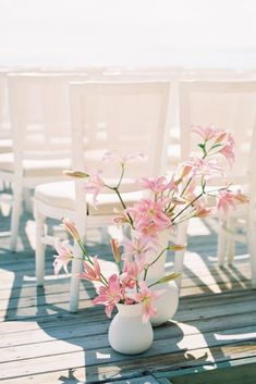 pink flowers in white vases sitting on a wooden deck next to chairs and the ocean