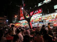 many people are sitting at tables in front of a food court with neon lights on the building