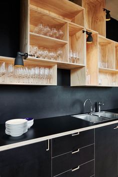 a kitchen with black counter tops and wooden shelves filled with wine glasses, plates and bowls