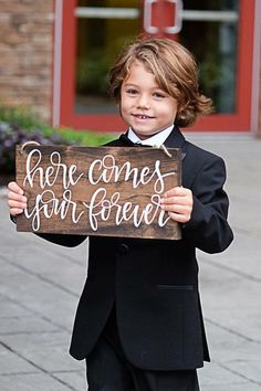 a young boy in a tuxedo holding a sign that says, here comes our forever