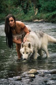 a woman in native garb standing next to a white wolf on a river bank