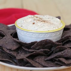 a white plate topped with a bowl of dip surrounded by tortilla chips on top of a wooden table