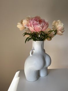 a white vase with pink flowers in it on a table next to a shadow cast wall