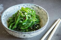 a white bowl filled with green vegetables next to chopsticks on a gray surface