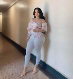 a woman standing next to a wall wearing white pants and a pink floral top with ruffle sleeves