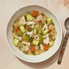 a bowl of chicken and vegetable soup next to a spoon on a white tablecloth