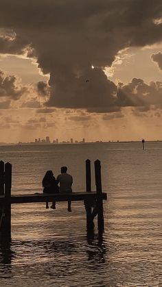 two people are sitting on a pier in the water under a cloudy sky at sunset