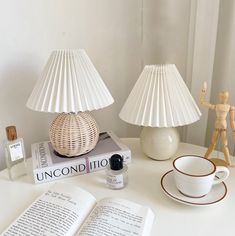 a white table topped with two lamps next to an open book and a cup on top of a saucer