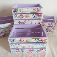 four boxes with floral designs on them sitting on a tablecloth covered surface, one is purple and the other has pink flowers