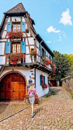 a woman walking down a cobblestone street past a house with flowers on the windows