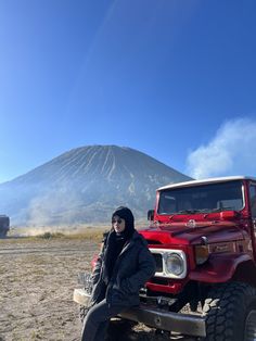 a woman is sitting on the hood of a red truck in front of a volcano