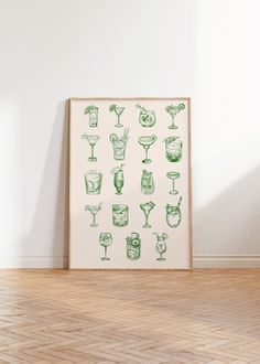 a framed poster with green cocktail glasses on it in front of a white wall and wooden floor