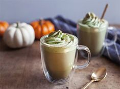 two cups filled with green tea and whipped cream on top of a wooden table next to pumpkins