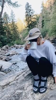 a woman sitting on top of a rock next to a river holding a coffee cup