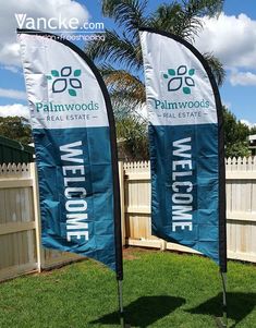 two blue and white welcome banners in the grass next to a fence with palm trees